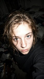 Grainy early-2000s style flash selfie of a young woman taken from above in a dim messy bedroom